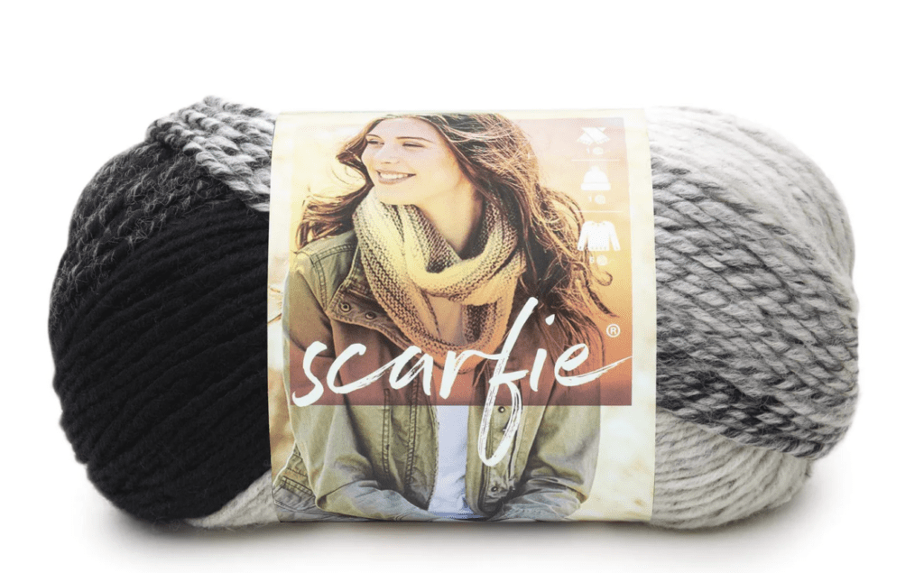 A skein of Scarfie yarn in a gradient of black, gray, and white. The label features a smiling woman wearing a knitted scarf and jacket, indicating the yarn's brand and potential use in creating wearable items. -Marly Bird